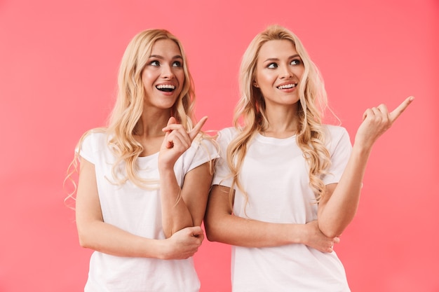 Uśmiechnięte bliźniaczki blond ubrane w t-shirty, wskazujące i odwracające wzrok na różowej ścianie