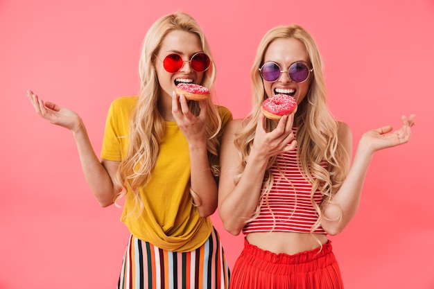 Uśmiechnięte blond bliźniaki w okularach przeciwsłonecznych jedzą razem pączki i na różowej ścianie