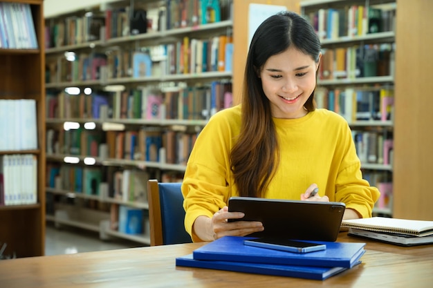 Uśmiechnięte studentki przygotowujące się do egzaminu, szukające informacji na tablecie cyfrowym w bibliotece