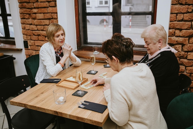 Uśmiechnięte trzy starsze ciemnowłose i jasnowłose kobiety w czarno-białych kurtkach grające w domino w kawiarni z przyjaciółmi celebrujące spotkanie przy stole z kieliszkami wina lub szampana na nim