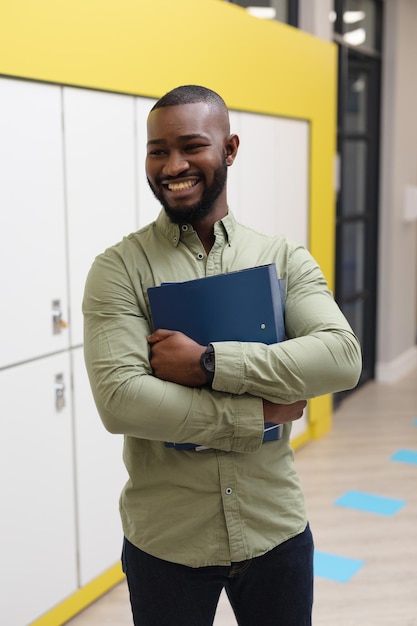Uśmiechnięty afroamerykański młody nauczyciel z plikami patrzący w stronę, stojąc na korytarzu