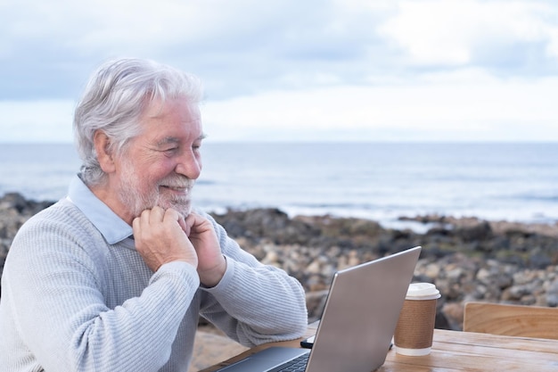 Uśmiechnięty atrakcyjny dojrzały starszy dorosły profesjonalny człowiek biznesu w zdalnej pracy na laptopie przy plaży Starszy brodaty mężczyzna siedzący przy drewnianym stole patrzący na ekran komputera