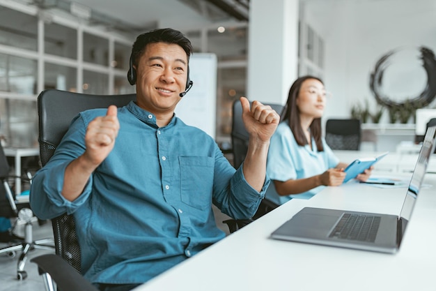 Uśmiechnięty azjatycki biznesmen pracujący na laptopie siedzący w coworkingu na tle kolegów