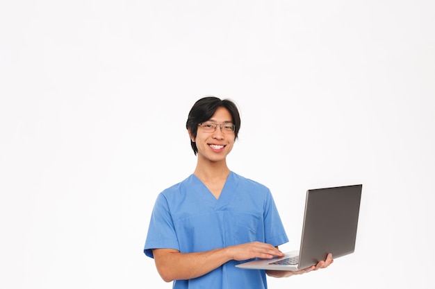 Uśmiechnięty azjatycki lekarz człowiek ubrany w mundur na białym tle nad białą ścianą, za pomocą komputera przenośnego