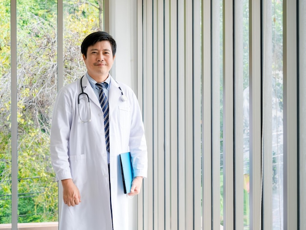 Uśmiechnięty azjatycki mężczyzna lekarz portret stojący w pobliżu szklanych okien w gabinecie medycznym z zielonym widokiem Pewny azjatycki dorosły mężczyzna lekarz ze stetoskopem trzymający niebieski folder z dokumentami patrząc na kamerę