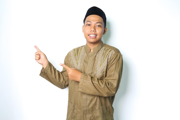 uśmiechnięty azjatycki muzułmanin wskazujący na prawą stronę, ubrany w islamską sukienkę odizolowaną na białym tle