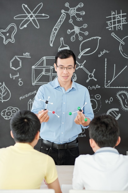 Uśmiechnięty azjatycki nauczyciel przedmiotów ścisłych, pokazujący ciekawym szkolnym molekularnym plastikowe modele