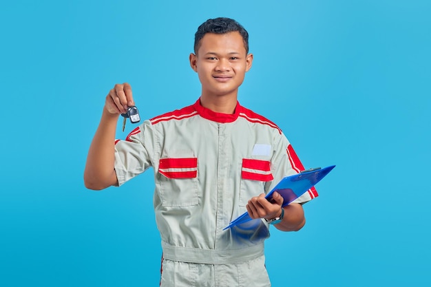 Uśmiechnięty azjatycki przystojny mężczyzna ubrany w mundur mechanika, trzymający schowek i pokazujący kluczyki do pojazdu
