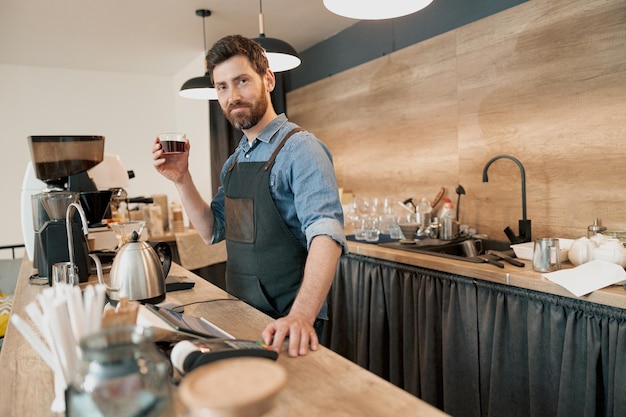 Uśmiechnięty barista trzymający w ręku szklankę z filtrowaną kawą i patrzący w kamerę