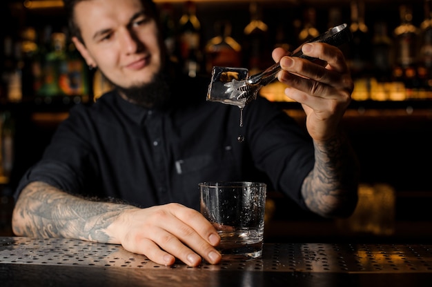 Uśmiechnięty barman dorzucający do drinka w szklance dużą kostkę lodu za pomocą pęsety na blacie barowym