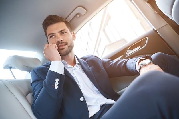 Uśmiechnięty biznesmen rozmawiający na smartfonie i siedzący na tylnym siedzeniu samochodu