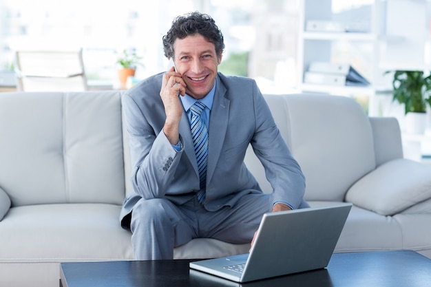 Uśmiechnięty biznesmen używa laptop i telefon komórkowego