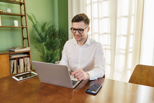 Uśmiechnięty biznesmen w białej koszuli pracuje na laptopie z tabletem i smartfonem na stole