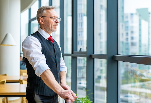 Zdjęcie uśmiechnięty biznesmen w szarym garniturze stojący i patrzący w okno dorosły mężczyzna w białej koszuli z okularami