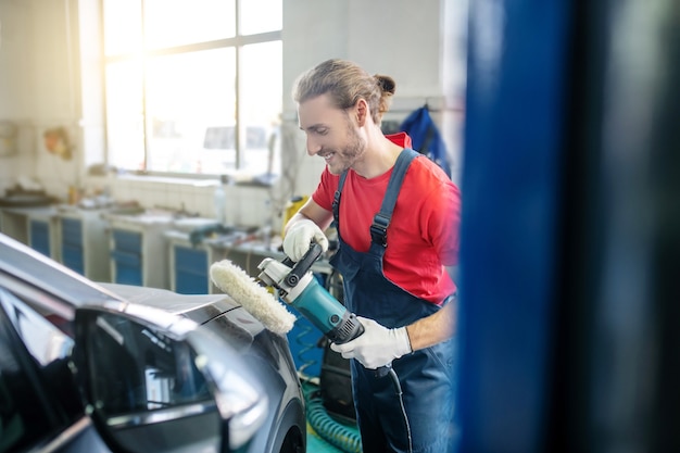 Uśmiechnięty brodaty mechanik w rękawicach ochronnych szlifowania karoserii w garażu