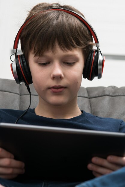 Uśmiechnięty brunetka mały chłopiec w przewodowych słuchawkach ogląda filmy słucha muzyki z cyfrowym tabletem w rękach siedzi na kanapie