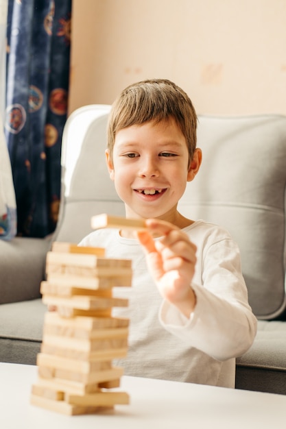 Uśmiechnięty Chłopiec Rasy Kaukaskiej Bawi Się W Domu, Na Stole W Jenga. Gry Planszowe Dla Dzieci I Dorosłych. Rozrywka Bez Gadżetów. Gry Podczas Wakacji W Domu. Szczęśliwe Dziecko Bawi Się Drewnianymi Klockami.