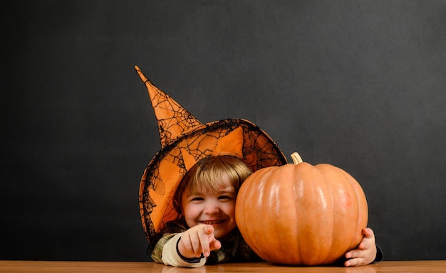 Uśmiechnięty chłopiec w kapeluszu wiedźmy z dynią halloweenową wskazującą na ciebie oszukać lub potraktować dziecko z dynią