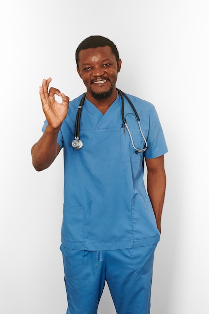 Uśmiechnięty czarny chirurg brodaty mężczyzna w niebieskim płaszczu ze stetoskopem robi OK gest