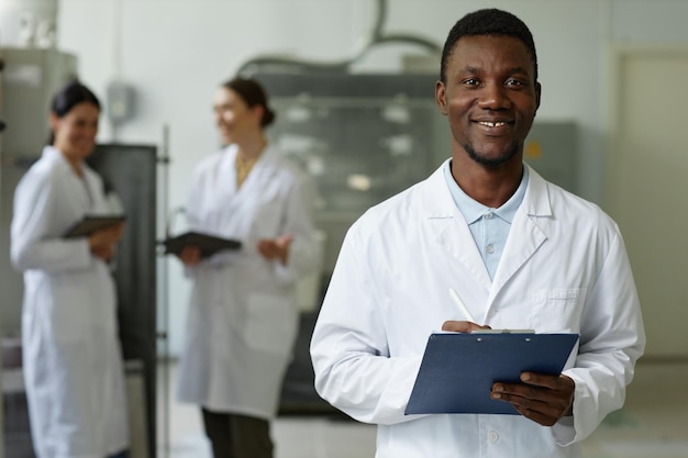 Uśmiechnięty czarny mężczyzna w płaszczu laboratoryjnym patrzący na kamerę w warsztacie fabrycznym