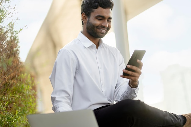 Uśmiechnięty Indianin trzymający telefon komórkowy za pomocą laptopa pracującego online na zewnątrz Udany biznes