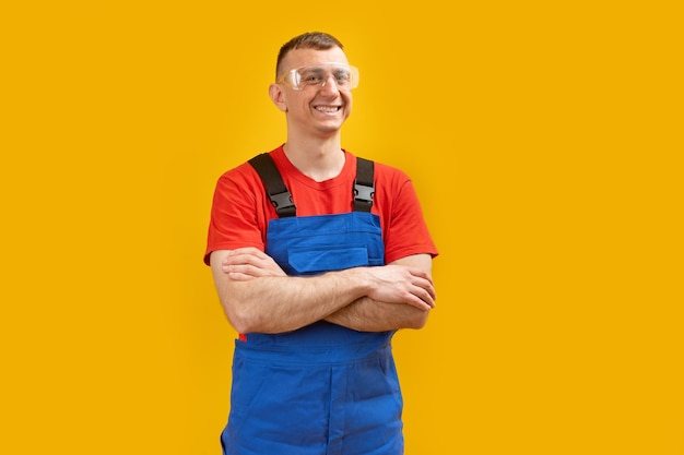 Uśmiechnięty inżynier w średnim wieku ubrany w niebieski kombinezon ze skrzyżowanymi ramionami stojący na białym tle na żółtym tle Kopiuj przestrzeń