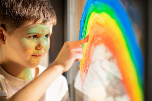 Zdjęcie uśmiechnięty ładny europejski mały dzieciak rysuje tęczę na oknie palcem i maluje z bliska rozbłysk słońca