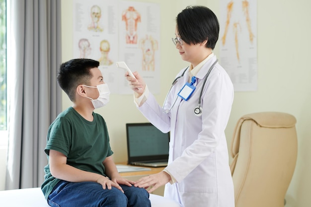 Uśmiechnięty lekarz sprawdzający temperaturę ciała chłopca za pomocą bezdotykowego termometru na podczerwień
