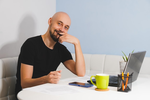 Uśmiechnięty łysy mężczyzna z brodą w czarnej koszulce pracuje nad swoim notebookiem w domu patrząc na kamerę