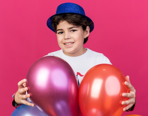 Uśmiechnięty Mały Chłopiec W Niebieskiej Imprezowej Czapce Stojący Za Balonami Odizolowanymi Na Różowej ścianie