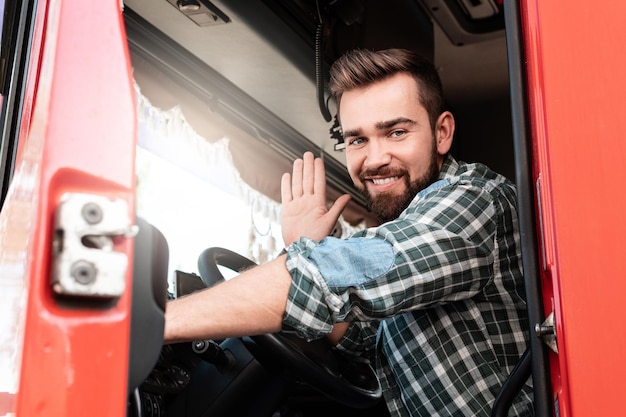 Uśmiechnięty mężczyzna kierowca ciężarówki siedzący w ciężarówce i machający ręką