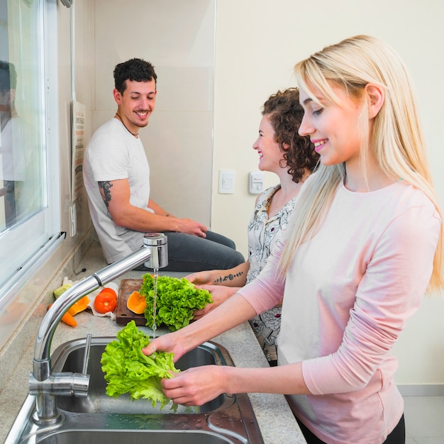 Zdjęcie uśmiechnięty mężczyzna obsiadanie na kuchennym worktop patrzeje dwa kobiety czyści sałaty