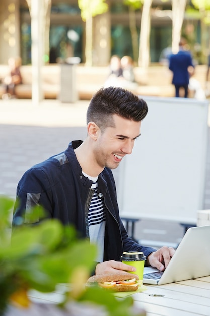 Uśmiechnięty mężczyzna pracuje na laptopie przy kawiarnią z kawą