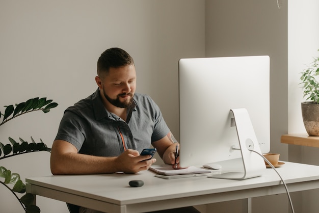 Uśmiechnięty Mężczyzna Pracuje Zdalnie Na Komputerze Stacjonarnym. Facet Z Brodą Robi Notatki I Trzyma W Domu Telefon Komórkowy. Nauczyciel Przygotowuje Się Do Wykładu Online.