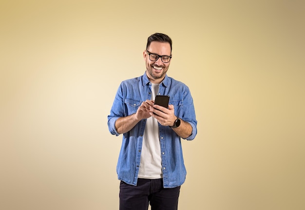 Uśmiechnięty mężczyzna przedsiębiorca ubrany w niebieską dżinsową koszulę wysyłający wiadomości tekstowe przez smartfon Szczęśliwy młody człowiek sprawdza media społecznościowe przez telefon komórkowy, stojąc odizolowany na beżowym tle