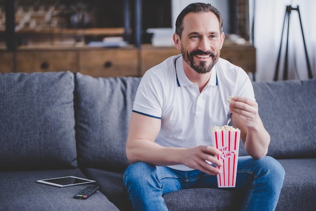 Uśmiechnięty mężczyzna siedzący na kanapie oglądając telewizję i jedzący popcorn w domu