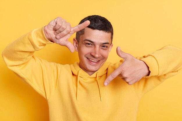 Uśmiechnięty mężczyzna ubrany w bluzę z kapturem w stylu casual, pozujący na białym tle na żółtym tle, patrzący na aparat przez palce gest ramki na zdjęcia, skupiający się i udając, że robi zdjęcie