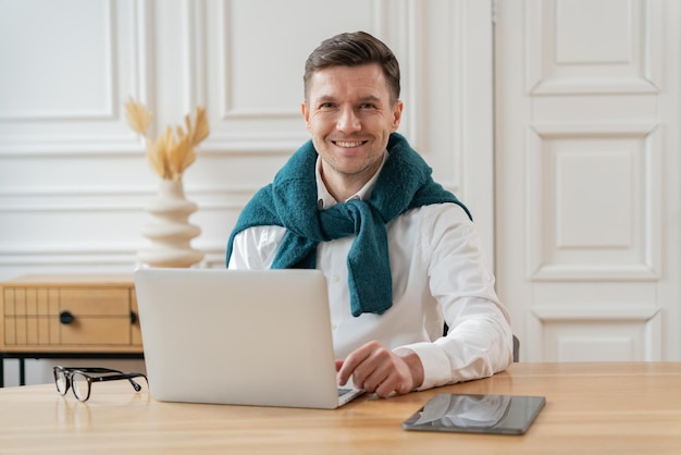 Uśmiechnięty mężczyzna w białej koszuli i błękitnym szalu pracuje na swoim laptopie przy eleganckim biurku z