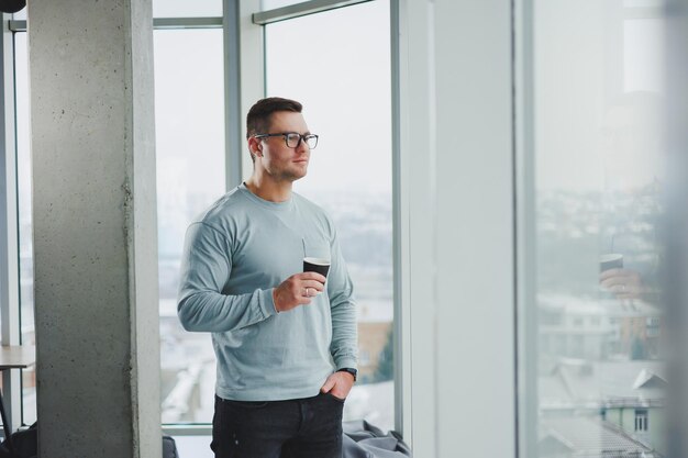 Uśmiechnięty mężczyzna w przypadkowych ubraniach stojący przy oknie z kawą w nowoczesnym miejscu pracy z dużym oknem w ciągu dnia