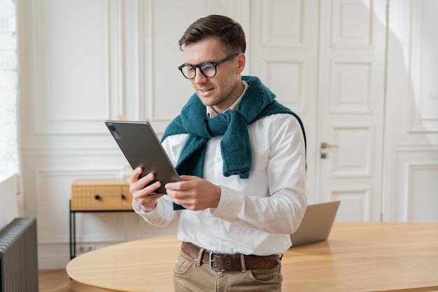 Uśmiechnięty mężczyzna z okularami używający tabletu przy biurku z laptopem i eleganckim dekorem na tle