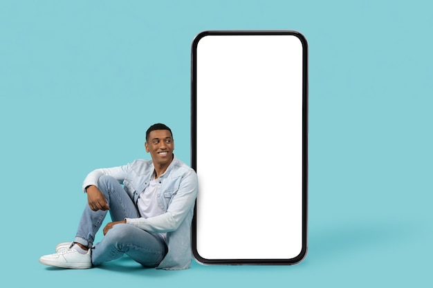 Uśmiechnięty młody Afroamerykanin w swobodnym siedzeniu na podłodze patrzy na ogromny smartfon z pustym ekranem