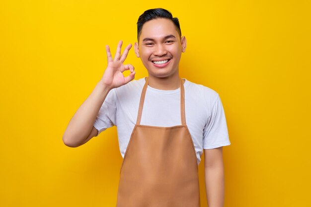 Uśmiechnięty młody Azjata, 20-letni pracownik baristy, ubrany w brązowy fartuch, pracujący w kawiarni, pokazujący dobry gest i najlepszą jakość lub dobrą ofertę na żółtym tle Koncepcja uruchomienia małej firmy