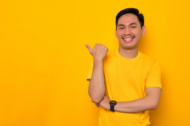 Uśmiechnięty młody Azjata w casualowej koszulce, wskazując kciukiem na bok na miejsce na białym tle na żółtym tle Świetna koncepcja oferty