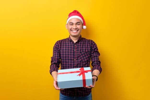 Uśmiechnięty młody Azjata w kapeluszu Mikołaja trzymający pudełko z wstążką na żółtym tle studia Szczęśliwego Nowego Roku 2023 celebracja wesołych świąt koncepcja