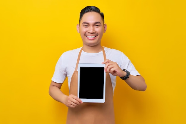 Uśmiechnięty młody azjatycki mężczyzna barista barman pracownik ubrany w brązowy fartuch pracujący w kawiarni pokazujący cyfrowy tablet z pustym ekranem do makiety izolowany na żółtym tle uruchamianie małej firmy