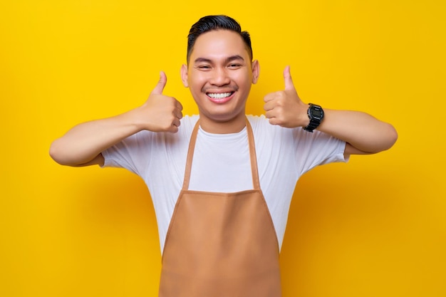 Uśmiechnięty młody azjatycki mężczyzna barista pracownik ubrany w brązowy fartuch pracujący w kawiarni Patrząc na kamerę pokazującą kciuki w aprobacie i polecającą na żółtym tle Koncepcja startowa małej firmy