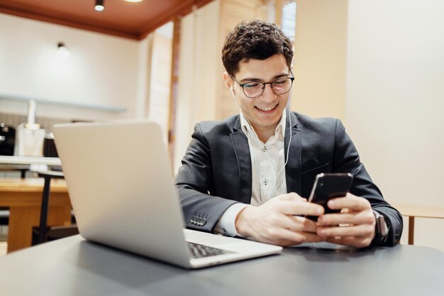 Uśmiechnięty młody biznesmen w garniturze używający smartfona i laptopa w kawiarni.