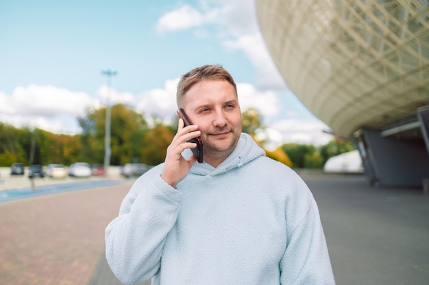 Uśmiechnięty młody brodaty biały bloger rozmawiający na smartfonie, stojąc na zewnątrz.