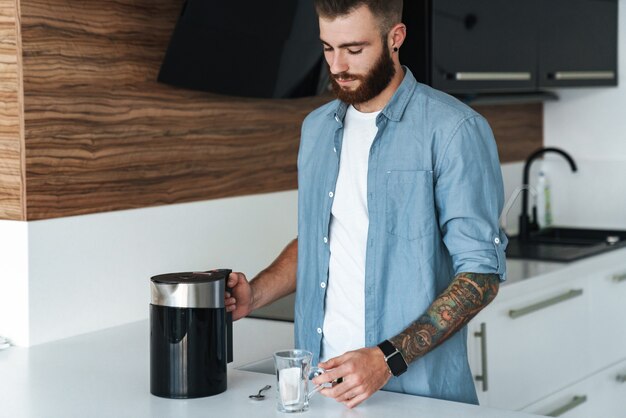 Uśmiechnięty młody brodaty mężczyzna robi filiżankę herbaty stojąc w kuchni w domu