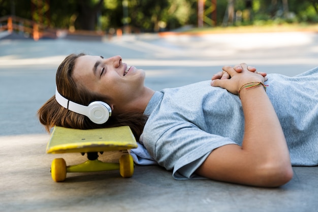 Uśmiechnięty młody chłopak spędza czas w skate parku, słuchając muzyki w słuchawkach, leżąc na deskorolce